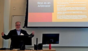 Arbitrator Pete Hanlon stands at a desk with arms outstretched. Behind him on a presentation screen is a page showing the title, "Keys as an Arbitrator."